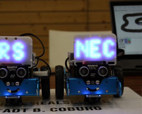 Zwei kleine ferngesteuerte Fahrzeuge mit nach vorne gerichteten Kameras und jeweils einer Platine, auf denen zusammen der Schriftzug RS NEC (staatliche Realschule Neustadt b. Coburg) zu erkennen ist.